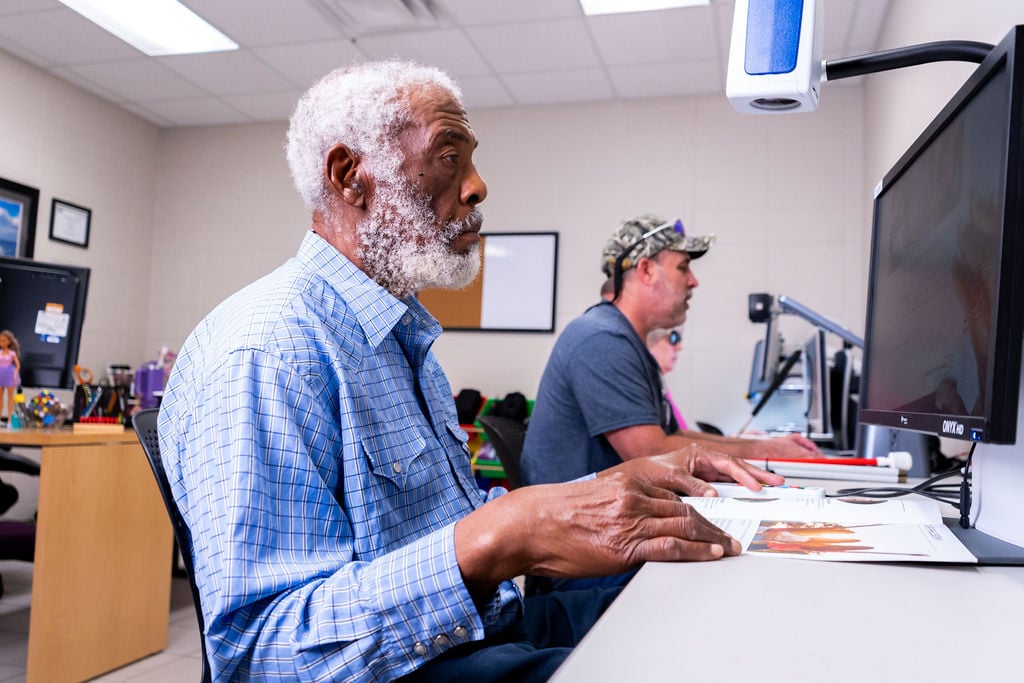 Older man with a gray beard typing at a computer in a shared workspace