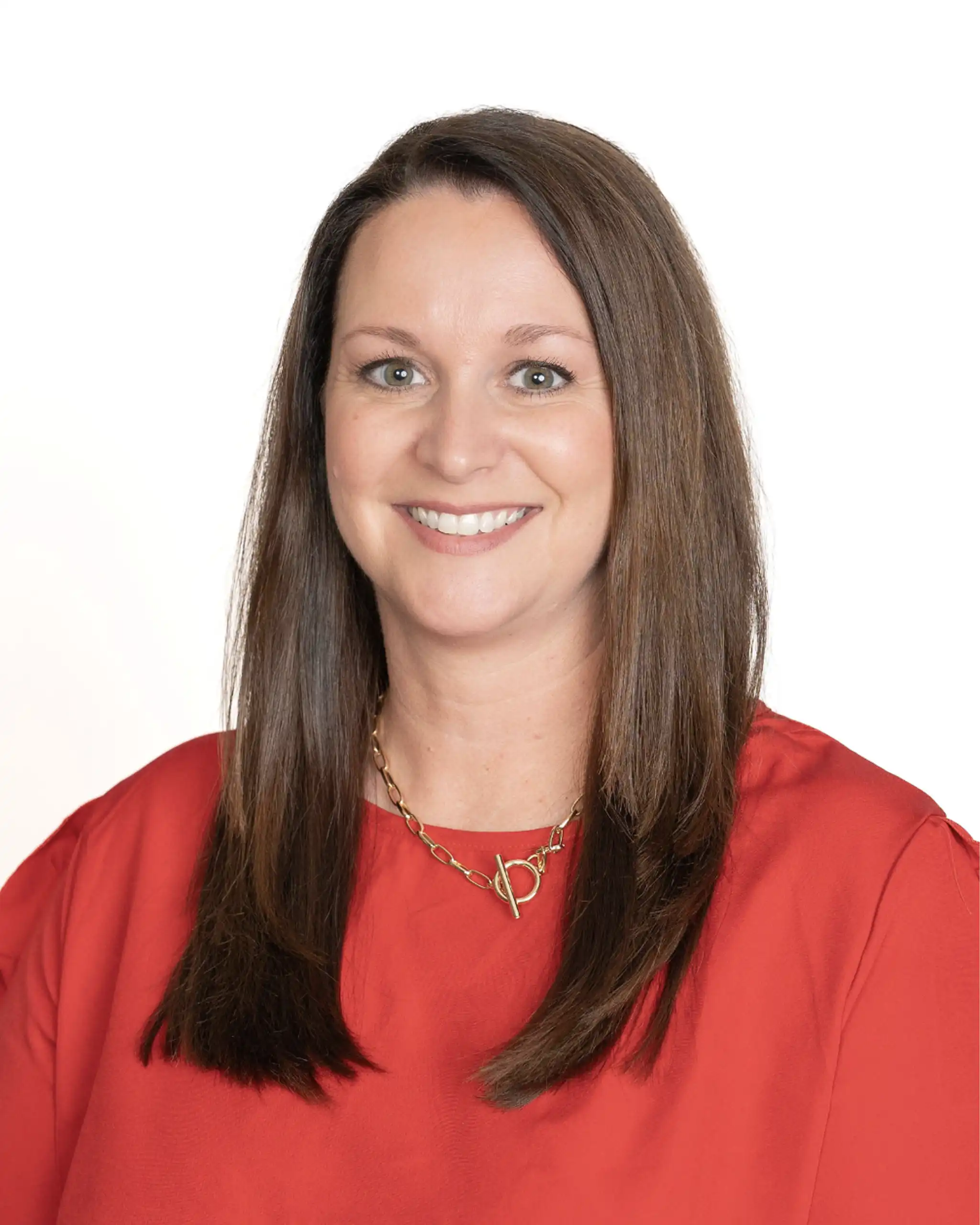 Smiling woman with long brown hair wearing a red blouse and gold necklace against a white background.