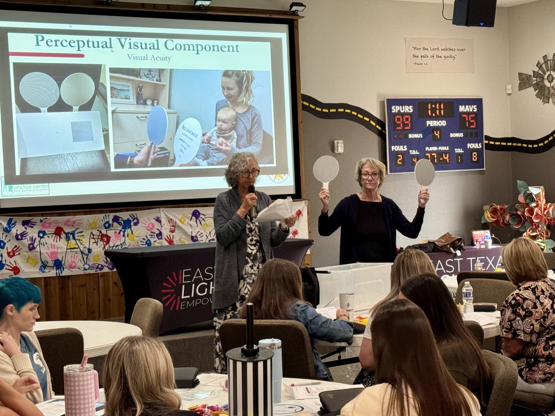 Two trainers stand at the front of a training room. One speaks into a microphone while the other holds up a pair of round gray acuity cards, one in each hand, at eye level. Behind them a projected slide reads “Perceptual Visual Component: Visual Acuity” and shows a parent holding an infant while a provider presents the cards. Participants watch from round tables in the foreground.