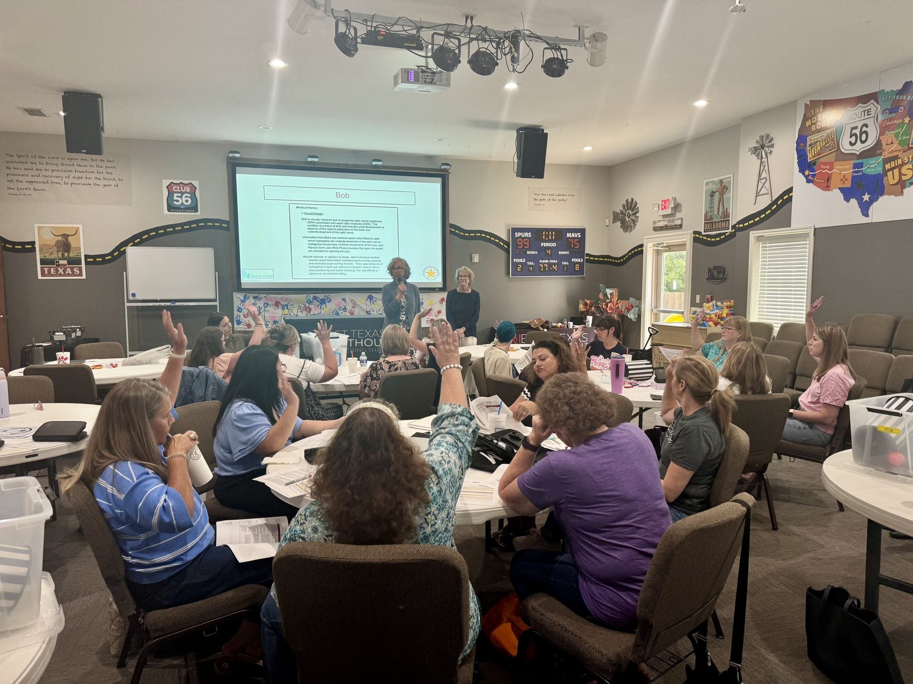 A wide view of the training room. About two dozen participants sit at round tables with printed packets, and several have a hand raised to answer a question. At the front, two trainers stand beside a projected case-study slide as they lead the discussion.