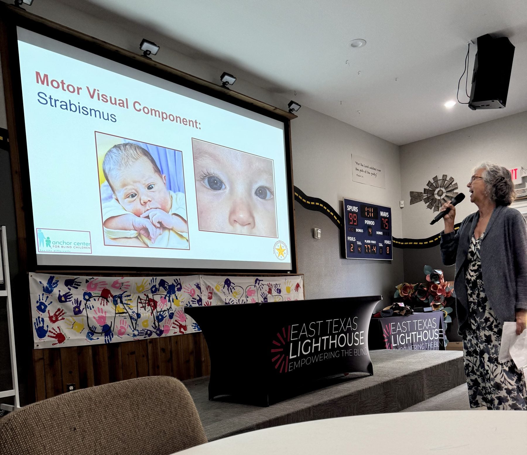 A trainer speaks into a microphone beside a projected slide titled “Motor Visual Component: Strabismus,” which shows two close-up photographs of an infant's eyes to illustrate eye alignment.